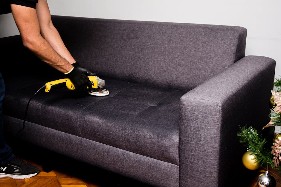 A person wearing black gloves is using a handheld electric fabric steamer or upholstery cleaning device to deep clean a dark grey fabric sofa in a residential living room. The sofa has a modern design