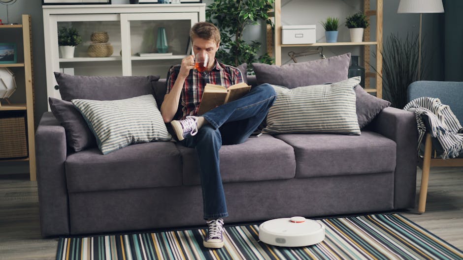 A person wearing black gloves is using a handheld electric fabric steamer or upholstery cleaning device to deep clean a dark grey fabric sofa in a residential living room. The sofa has a modern design