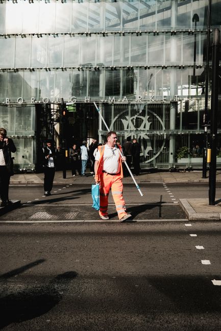 A commercial cleaning worker in an orange uniform and white shirt walking across a paved street outside the Boroondara Market building, which features a modern, glass-fronted exterior with reflective